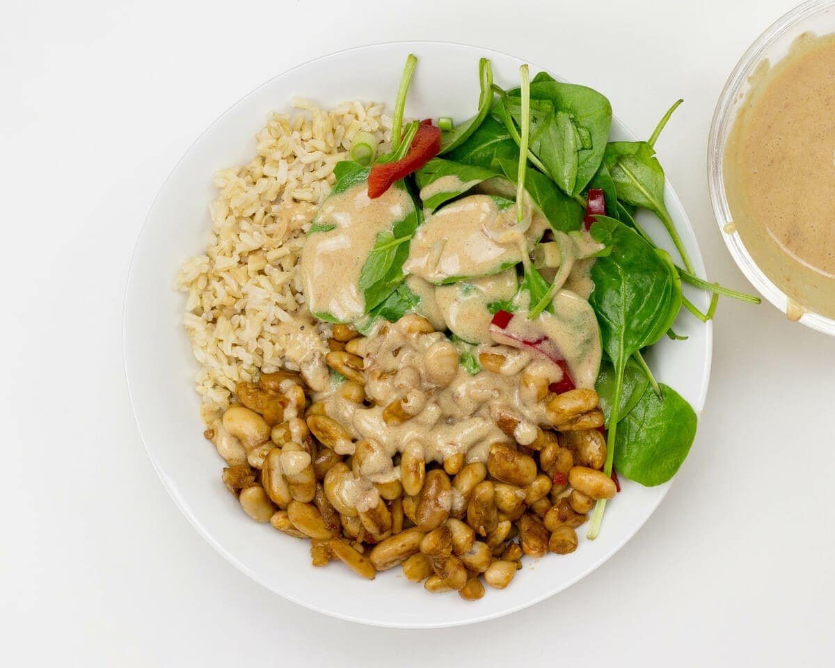 A white bowl containing brown rice, baked beans, and a spinach salad topped with a creamy dressing. A side container of additional dressing is partially visible on the right.