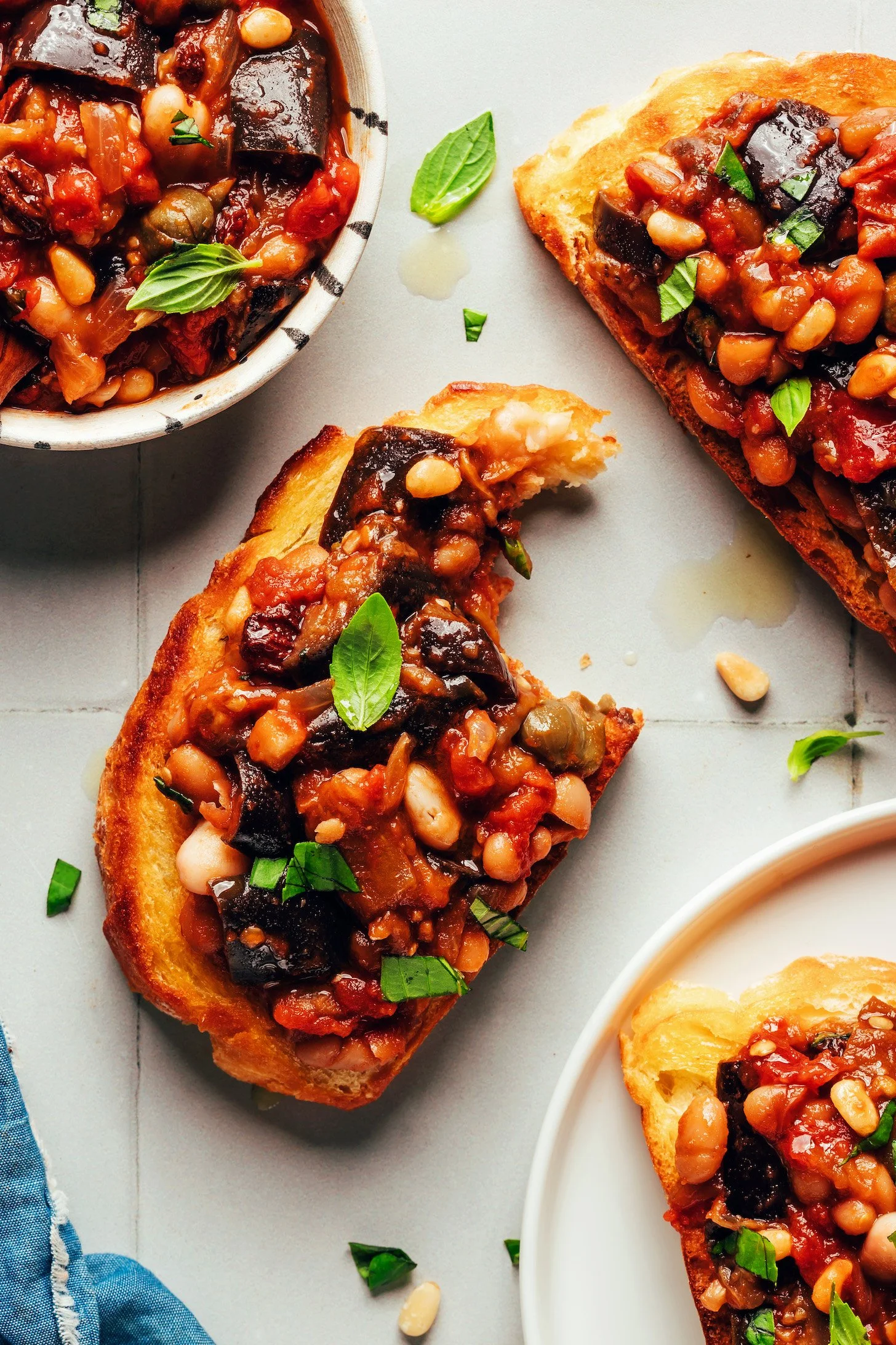 Close-up of toasted bread topped with a vegetable and bean stew garnished with fresh basil and chopped herbs, served on a white surface.