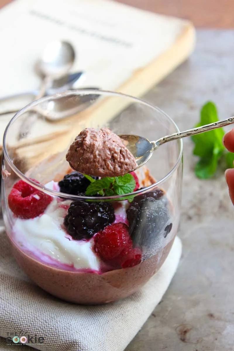 Close-up of a glass jar filled with chocolate mousse topped with mixed berries, a sprig of mint, and a spoonful of whipped topping, sitting on a beige cloth with a spoon and book in the background.