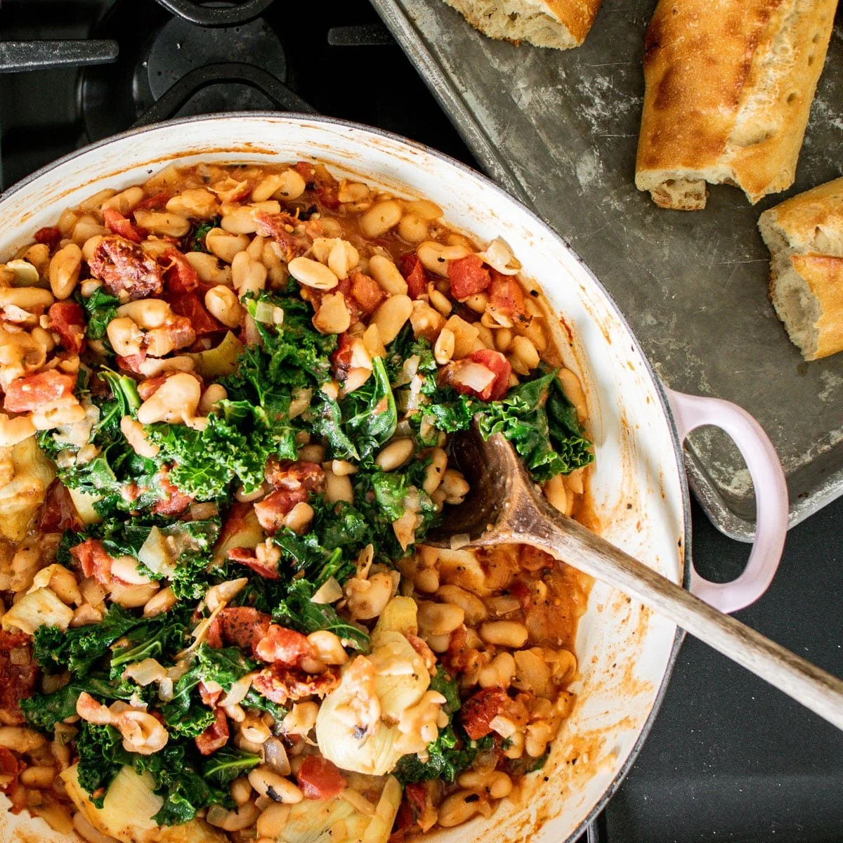 A white baking dish containing a bean, kale, and tomato stew, with a wooden spoon inside. There are pieces of torn bread on a tray nearby.