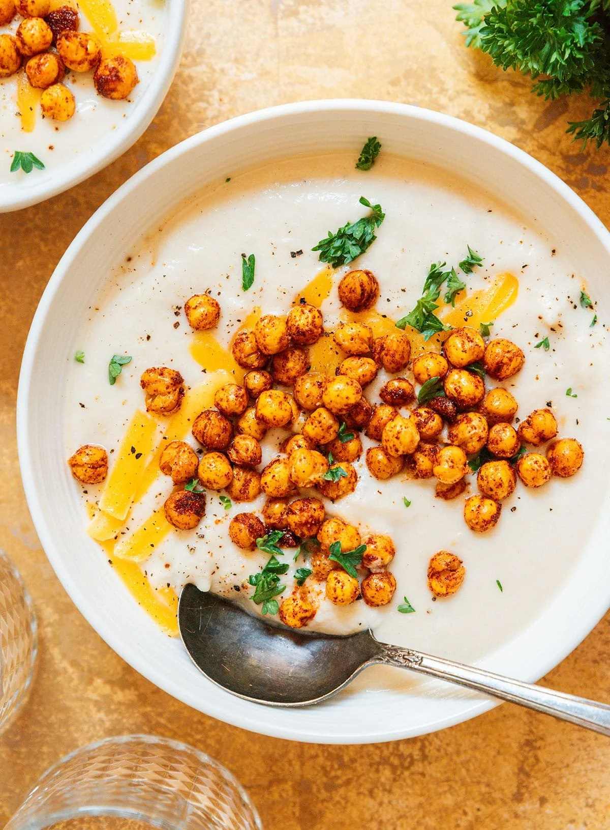 A bowl of creamy soup topped with roasted chickpeas, chopped parsley, and a drizzle of yellow sauce, with a spoon resting on the bowl.
