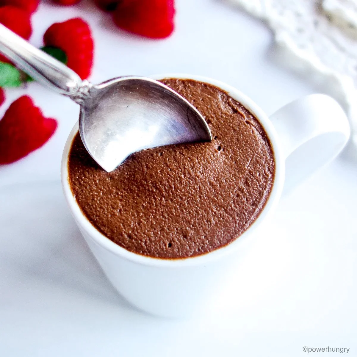 A white mug filled with vegan hot chocolate topped with a layer of foam, with a silver spoon inside, placed on a white surface with strawberries and whipped cream nearby.