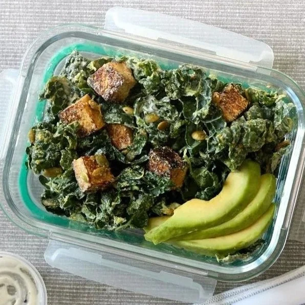 Kale salad with croutons and sunflower seeds, served with sliced avocado in a glass container.