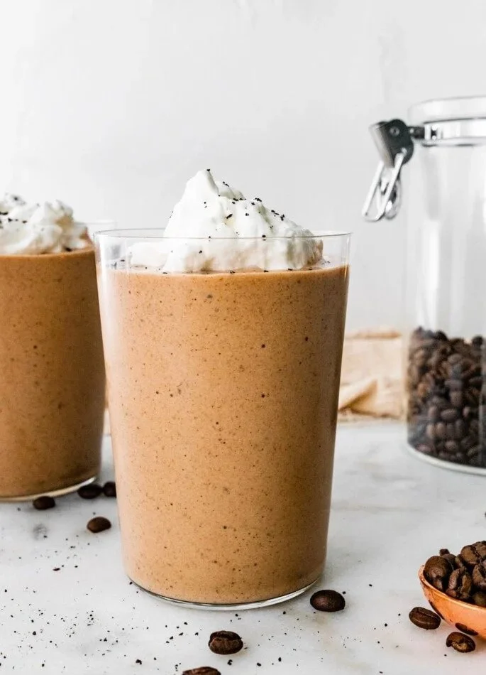 Glass of iced coffee topped with whipped cream surrounded by coffee beans and coffee containers on a white surface.