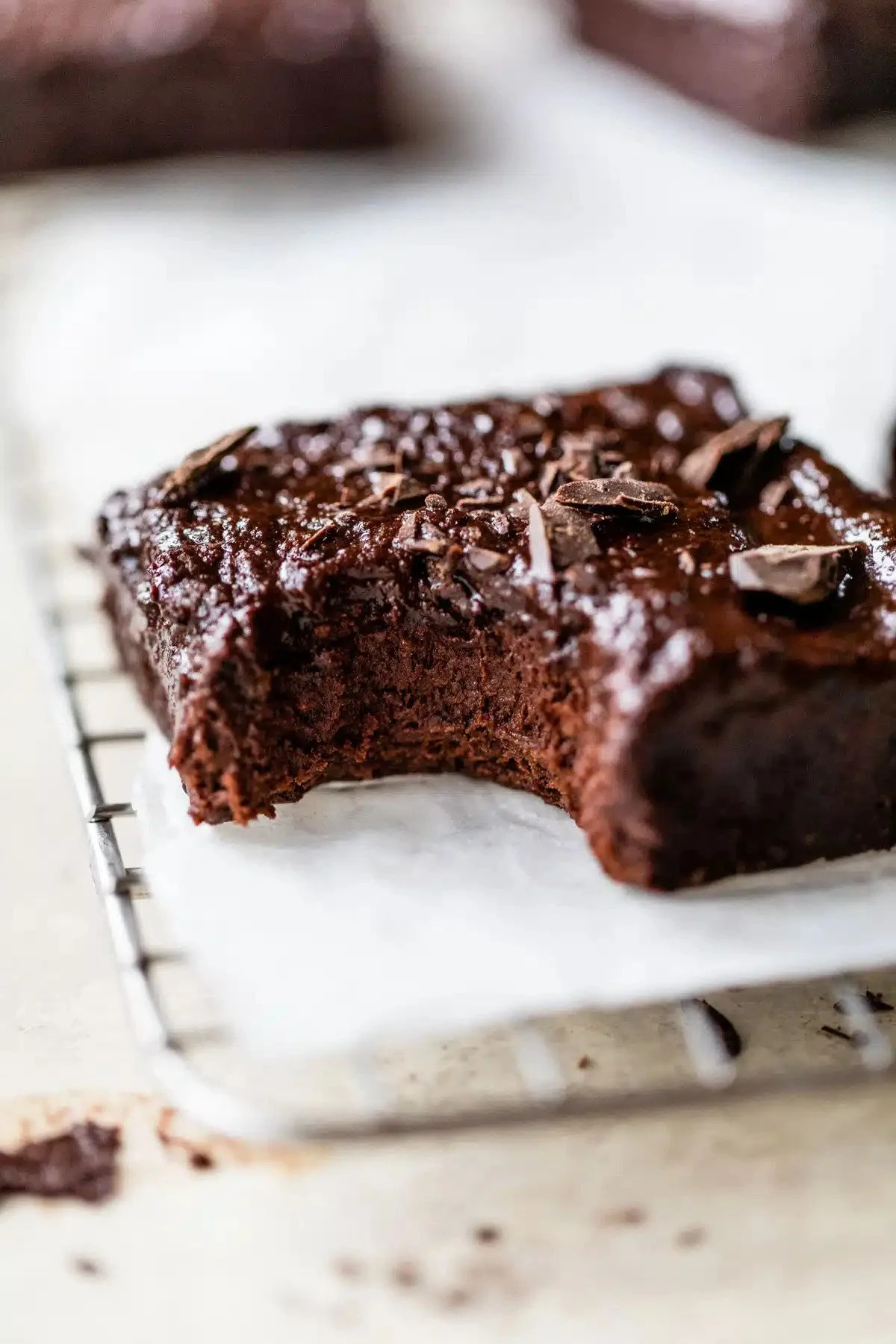 A partially eaten vegan chocolate brownie topped with chocolate shavings resting on parchment paper on a cooling rack.
