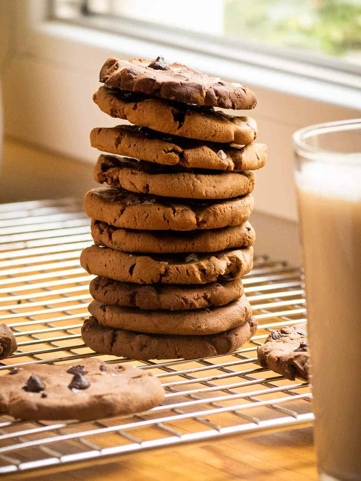 A tall stack of chocolate chip cookies on a cooling rack near a window with a glass of milk nearby.