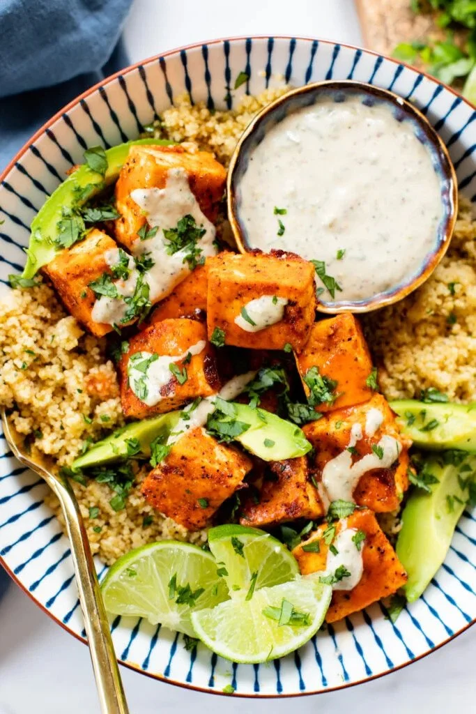 A bowl of chicken tikka served over couscous, garnished with fresh cilantro and lime wedges, with a side of creamy white sauce.