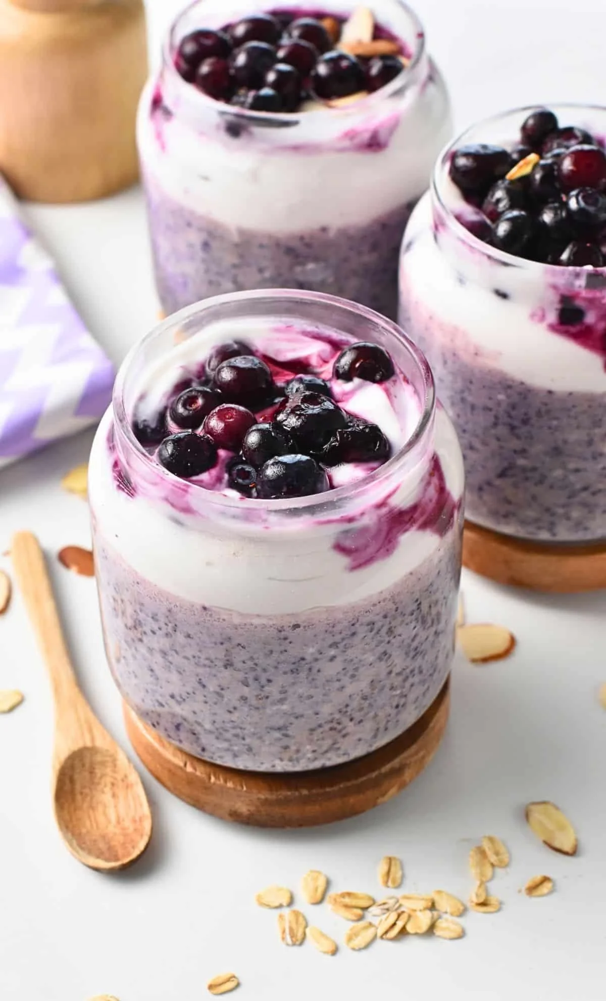 Three glasses of blueberry chia seed pudding topped with blueberries, served on wooden coasters with scattered oats and a wooden spoon nearby.