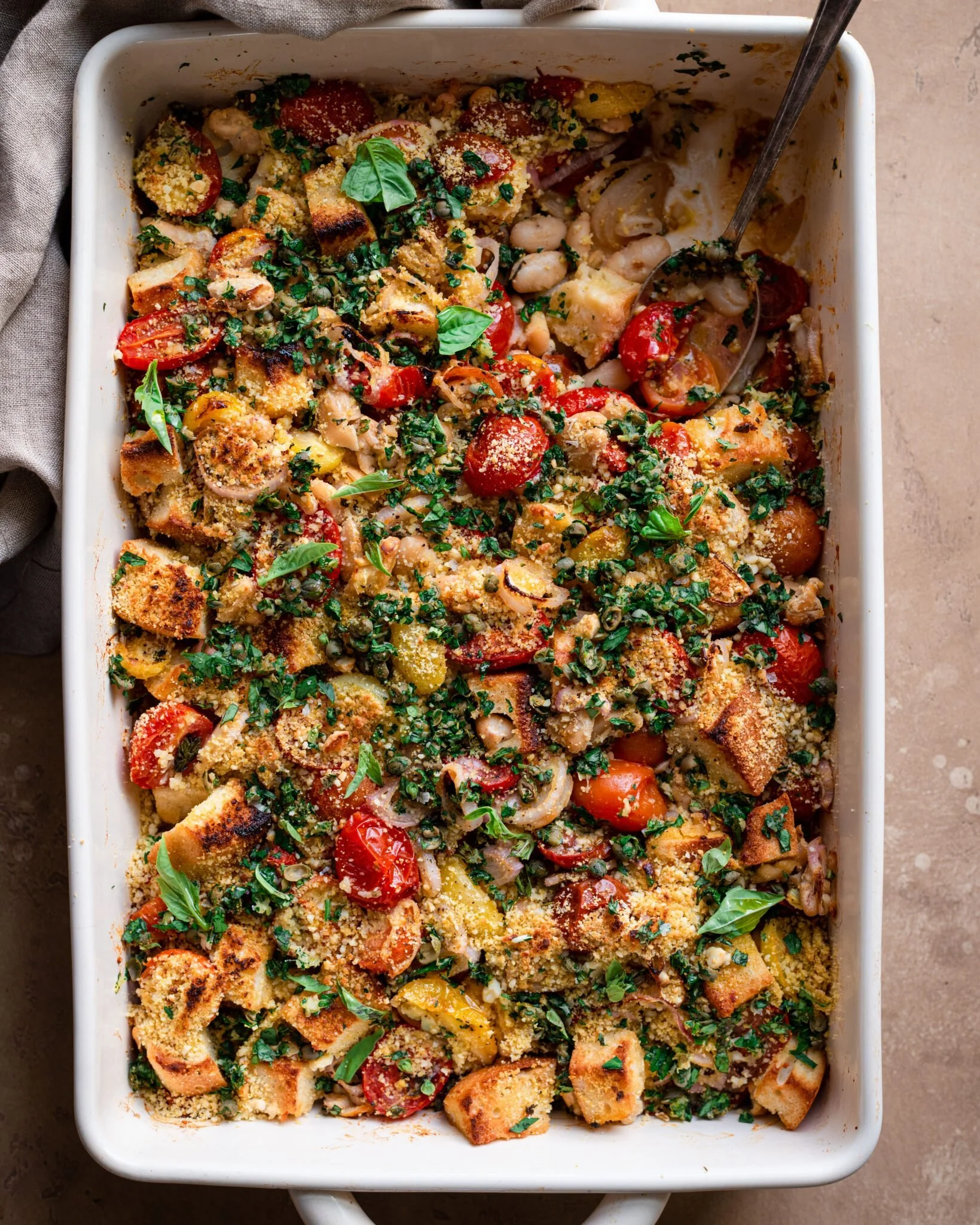 Baked casserole dish with cherry tomatoes, bread cubes, vegetables, herbs, and grated cheese, topped with fresh basil.