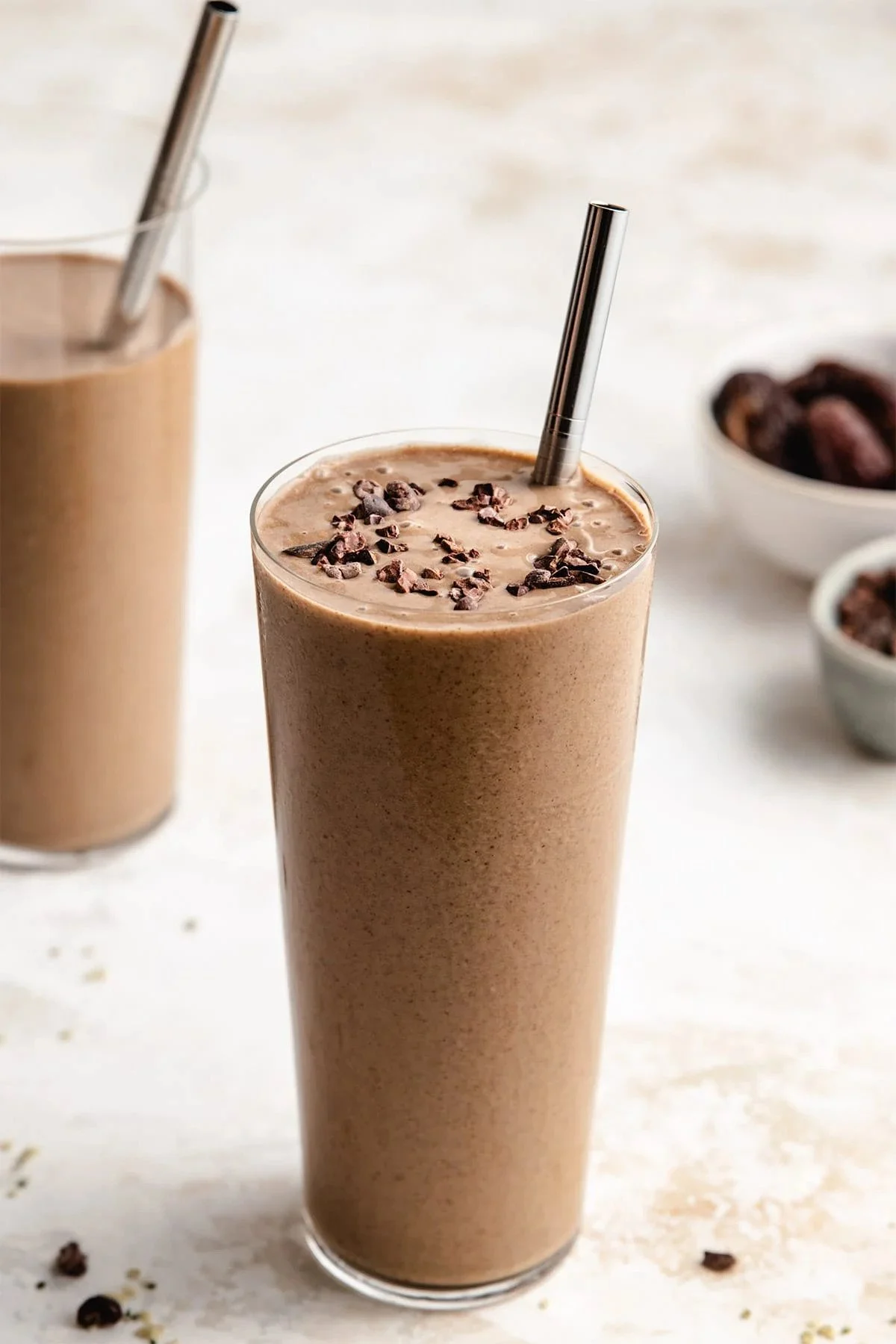 A tall glass of vegan chocolate smoothie topped with cocoa nibs and a metal straw on a light surface.