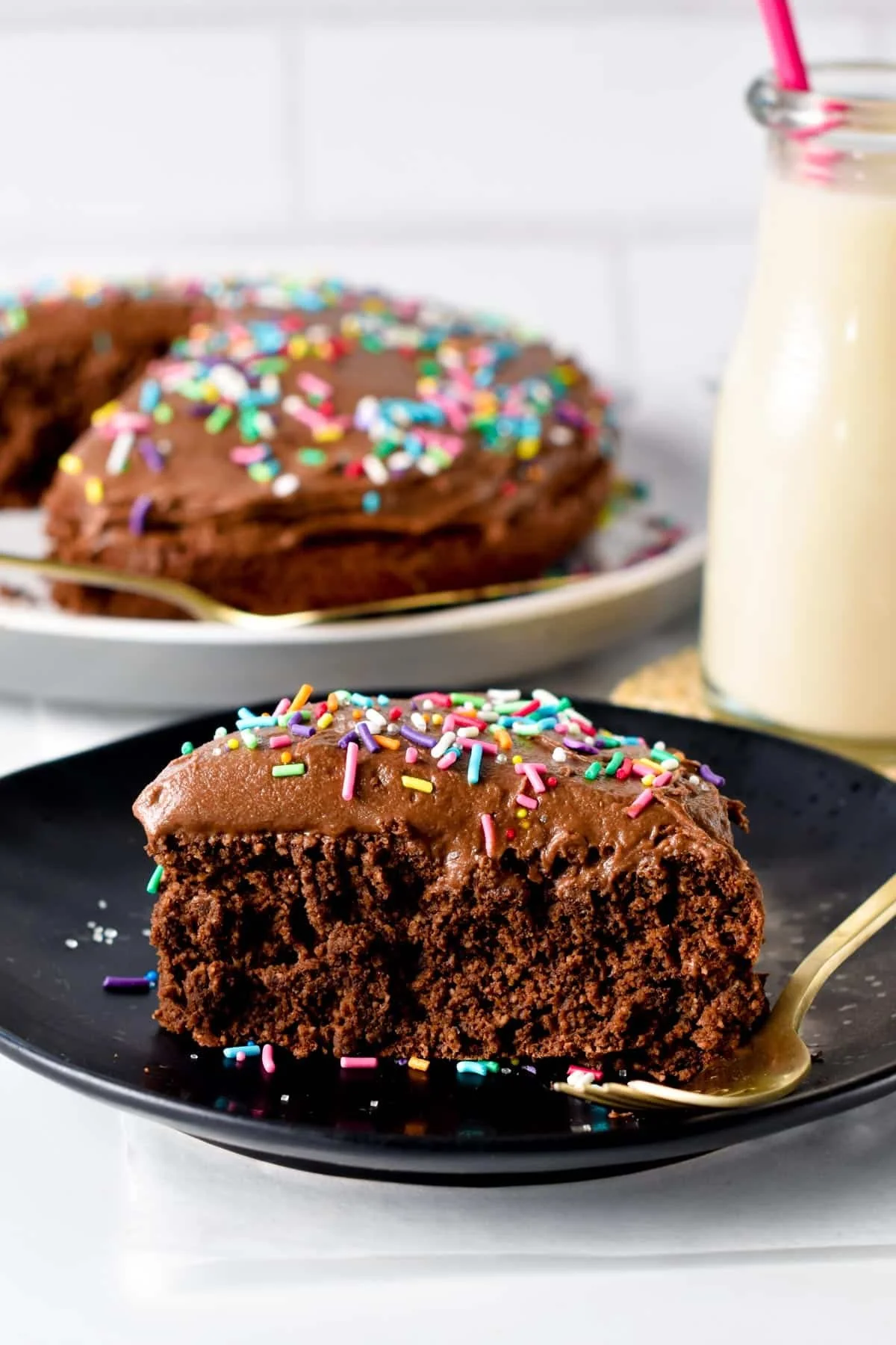 A slice of chocolate cake with colorful sprinkles on a black plate and an entire cake with sprinkles on a white plate in the background, with a jar of milk.