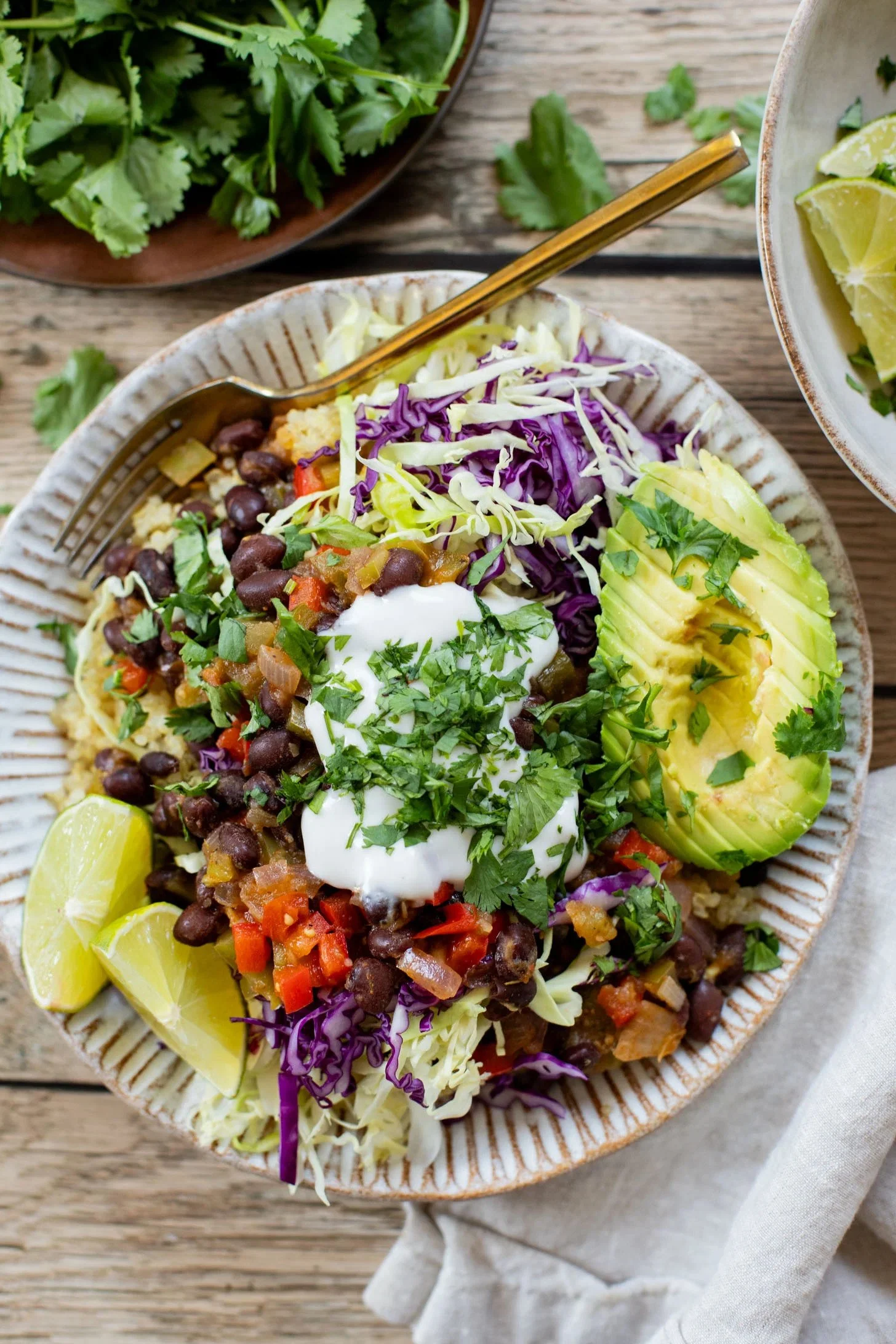 A plate of Mexican or Tex-Mex style salad with black beans, chopped vegetables, shredded purple cabbage, sliced avocado, lime wedges, sour cream, and chopped cilantro.