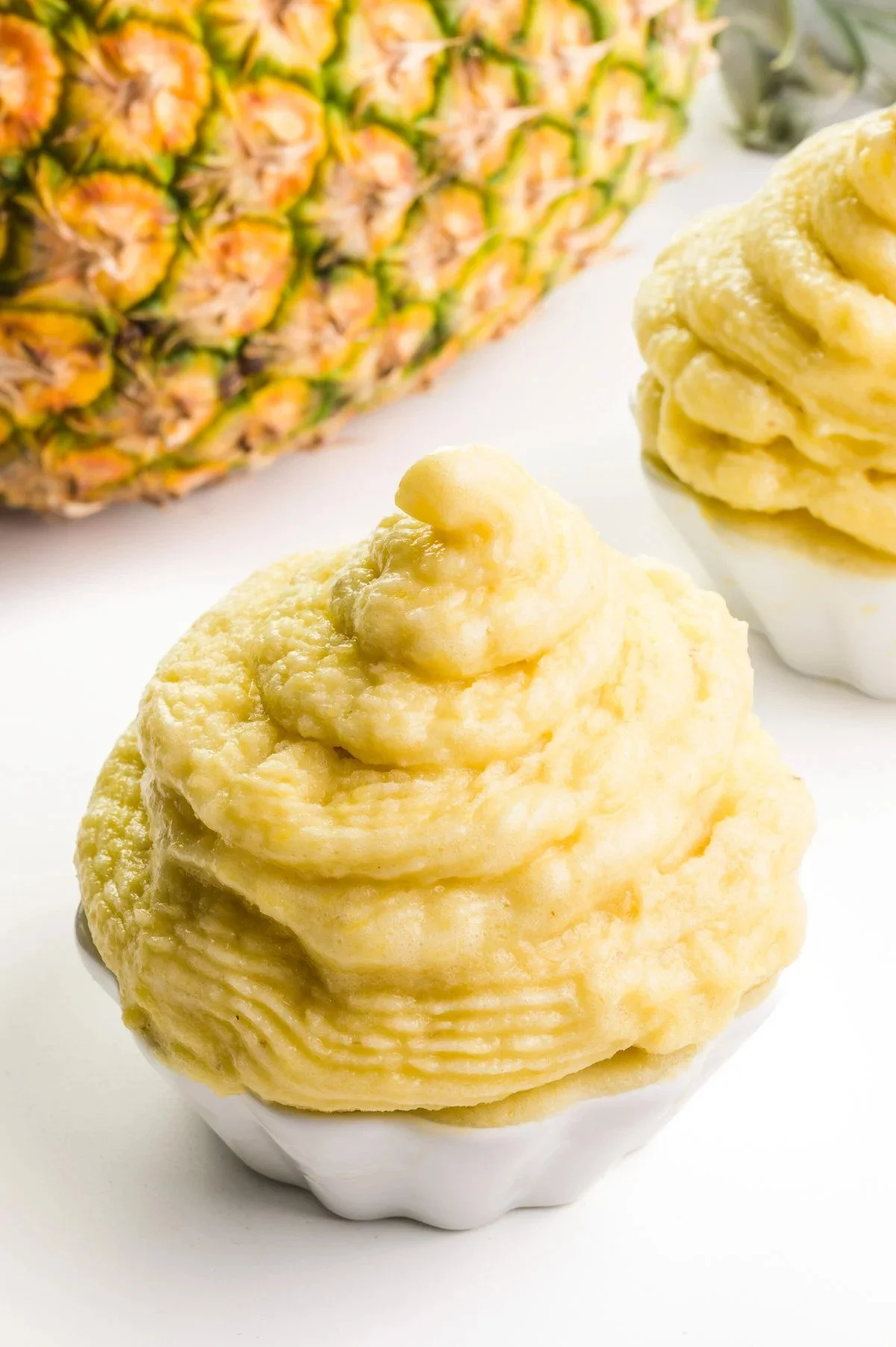 Pineapple-shaped frozen dessert with a creamy texture in a white cupcake liner, with a fresh pineapple in the background.