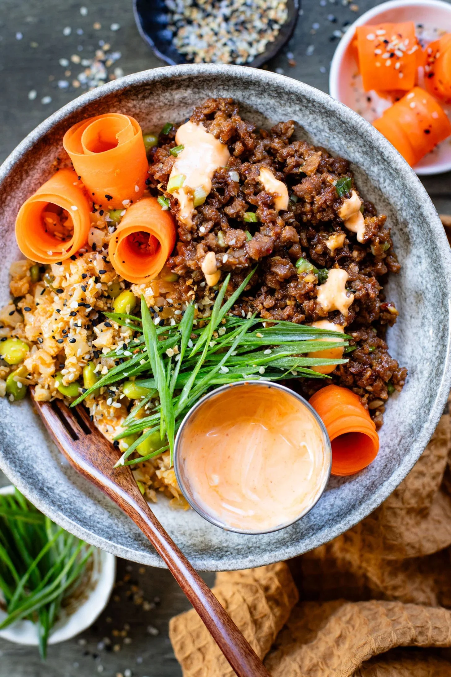 Vegan Power Bowl with orange carrot curls, chopped green onions, chopped nuts, and sesame seeds, accompanied by a small cup of sauce, with separate small plates of carrots and chopped vegetables, on a wooden table.