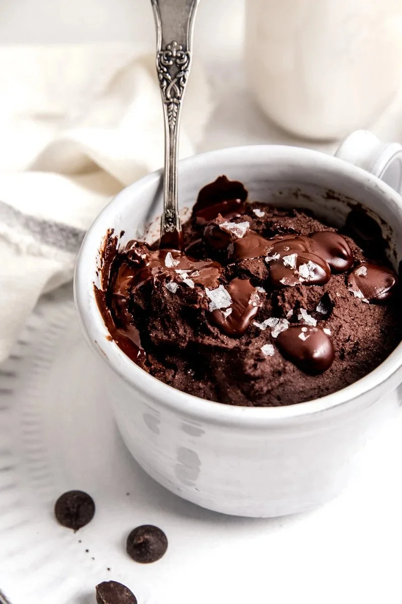 A white mug filled with melted chocolate brownie topped with chocolate chips and sea salt flakes, with a spoon inside. There are a few chocolate chips scattered on the white surface and a white cloth is in the background.