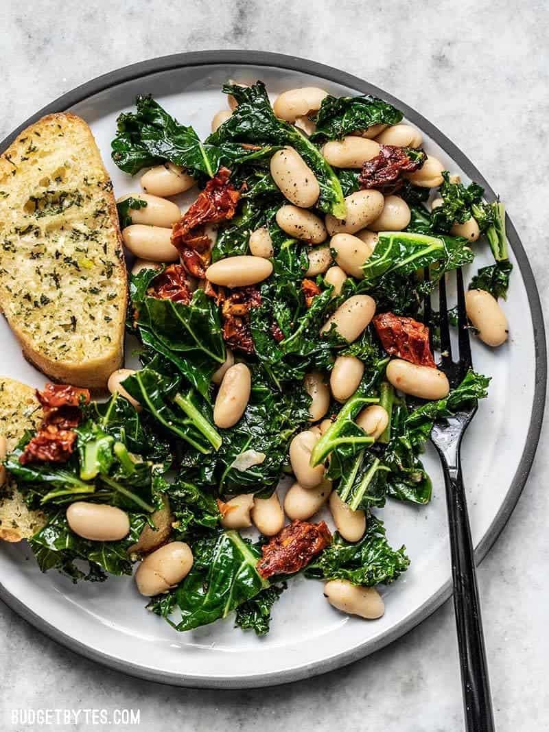 Kale salad with white beans and sun-dried tomatoes on a round plate with garlic bread on the side.