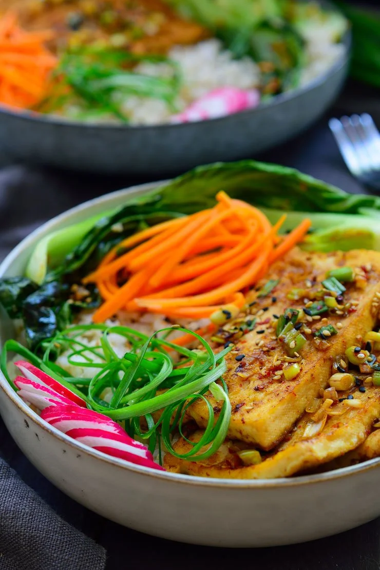 A bowl of Asian-style tofu with sliced radishes, spiralized green onions, and julienned carrots, garnished with chopped green onions and sesame seeds, with a side of leafy greens.
