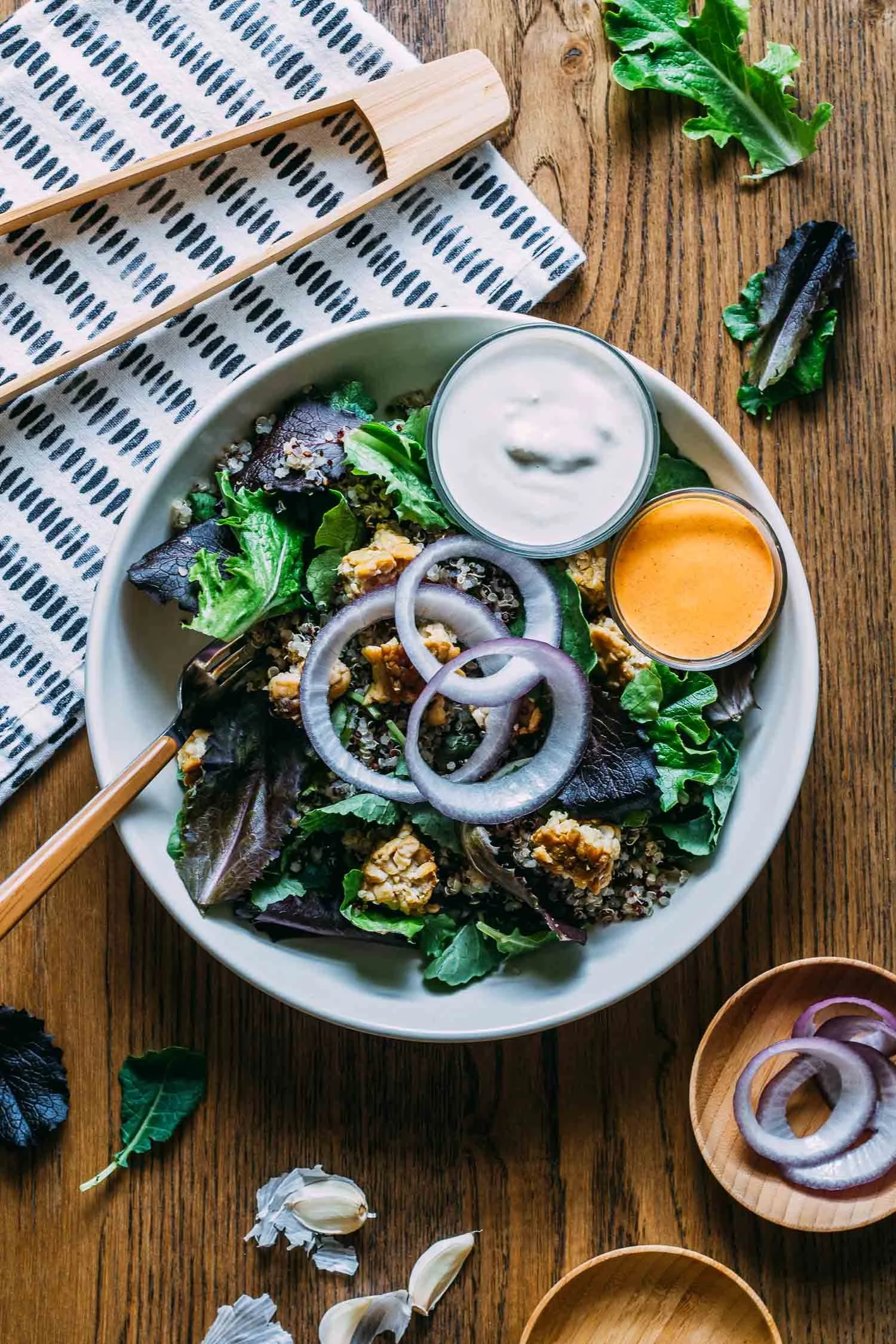 A bowl of mixed greens salad with red onion slices, quinoa, and two dressings, one creamy and one orange, on a wooden table, with garlic cloves, onion rings, and decorative chopsticks nearby.