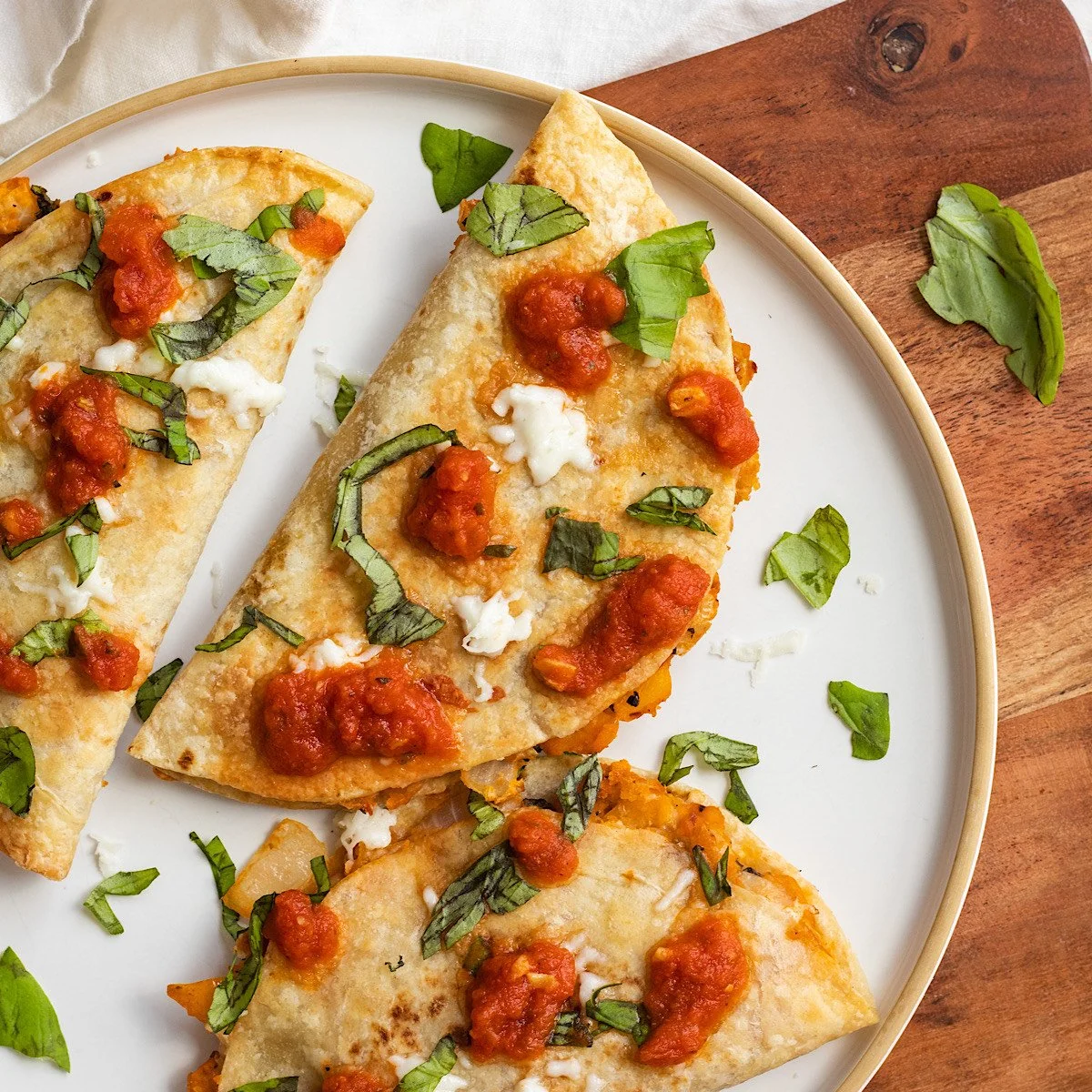 Close-up of three pizza slices topped with tomato sauce, melted cheese, and chopped basil leaves on a white plate with a wooden background.