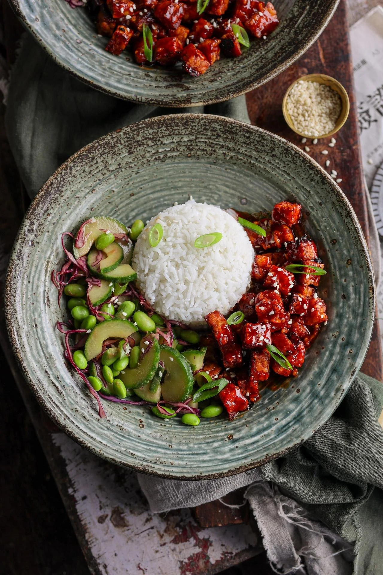 A bowl of Asian-style food with a mound of white rice, glazed tofu pieces garnished with green onion, and a side of cucumber salad with edamame and purple cabbage.