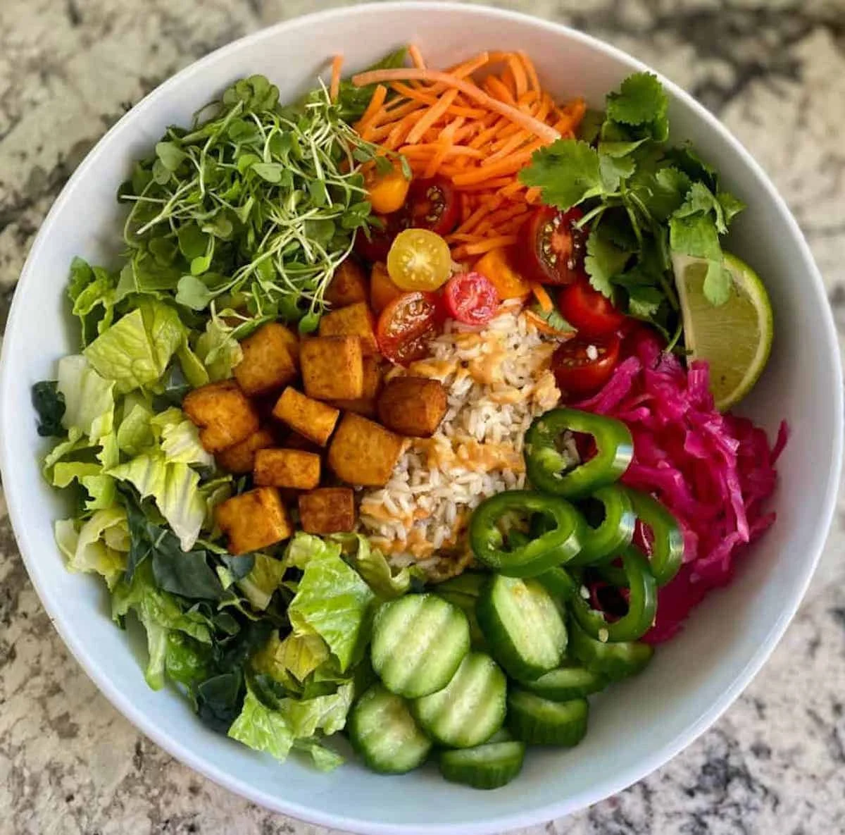 Colorful vegetarian bowl with fresh vegetables, tofu cubes, rice, cherry tomatoes, shredded carrots, cucumber slices, pickled cabbage, cilantro, lime wedge, and microgreens on top.