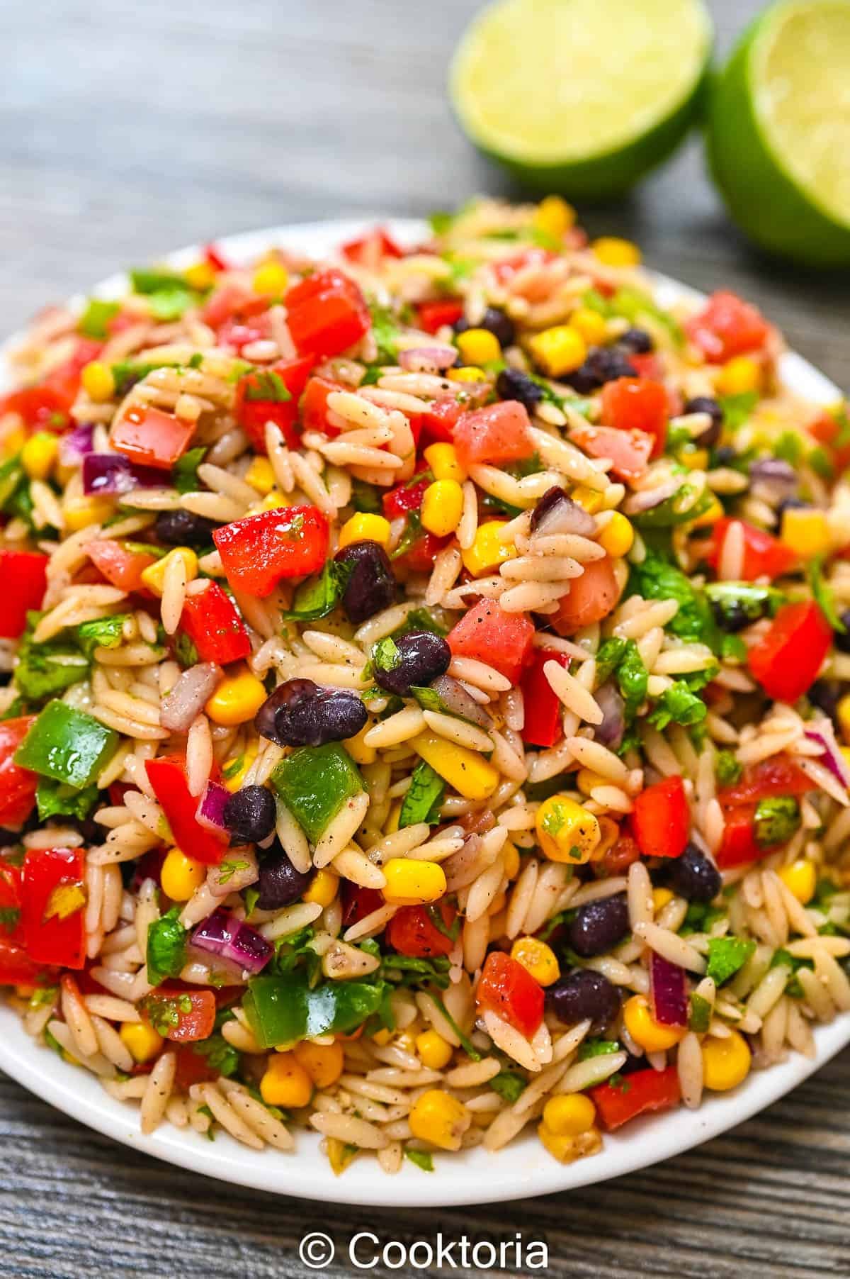 Colorful rice salad with diced red peppers, corn, black beans, green onions, and chopped herbs in a white bowl on a wooden surface with lime halves in the background.