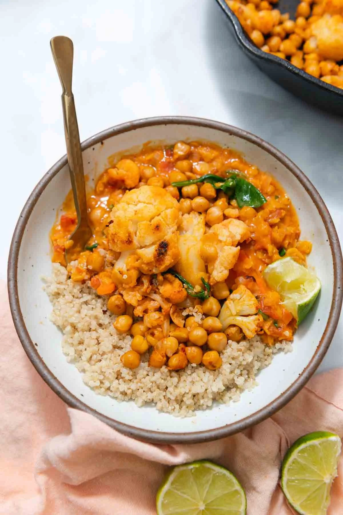 A bowl of chickpea and cauliflower curry with rice, topped with a lime wedge and a spoon, on a pink cloth, with more curry in a black dish in the background.