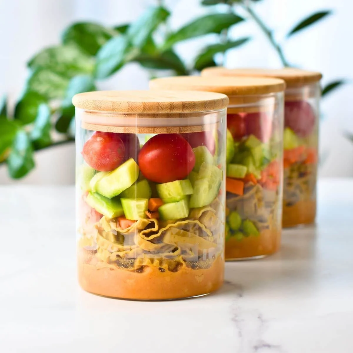Three glass jars filled with chopped vegetables, sealed with wooden lids, with a leafy green plant in the background.