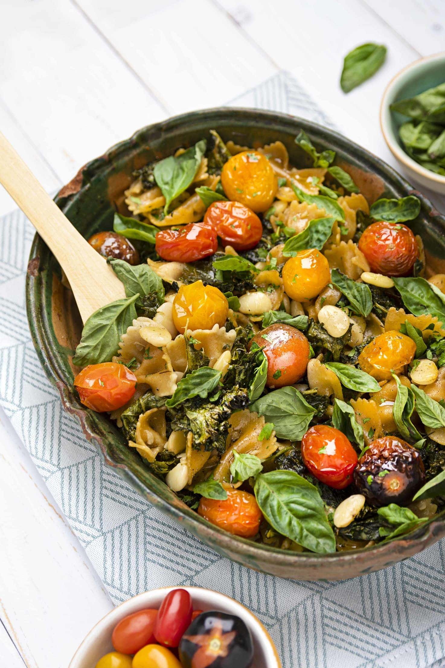 A bowl of roasted cherry tomatoes, pasta, and fresh basil leaves on a white tablecloth with a small bowl of cherry tomatoes in the foreground.