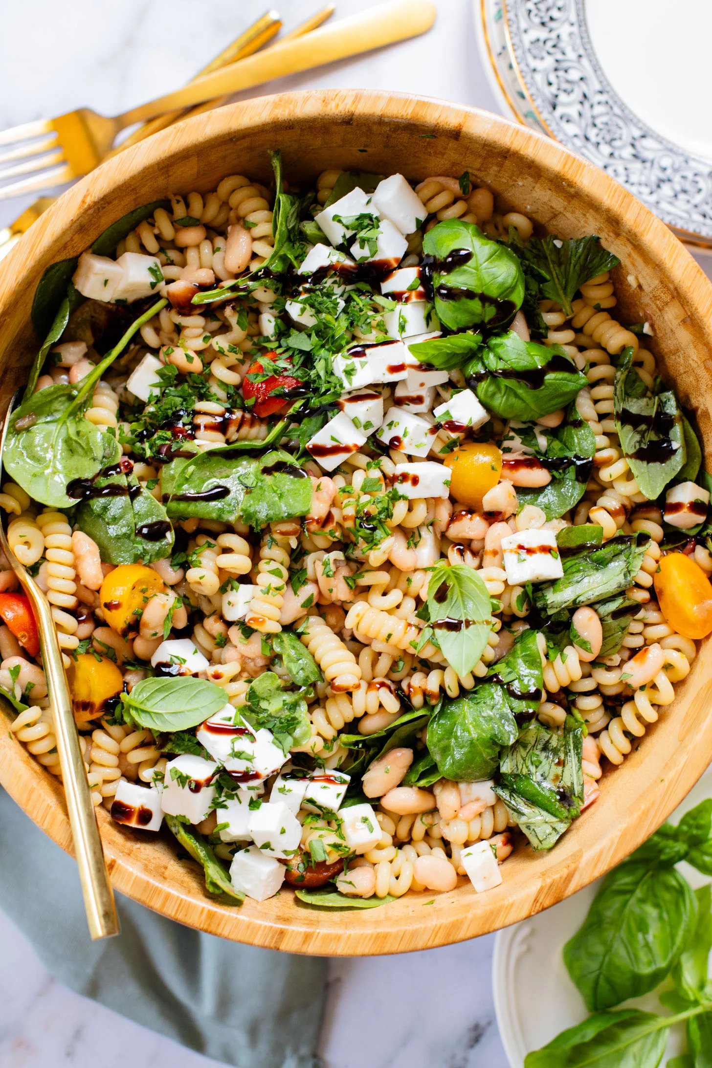 A bowl of pasta salad with fusilli pasta, cherry tomatoes, fresh basil and spinach, diced mozzarella cheese, chopped parsley, and balsamic glaze drizzle, served with a fork and a decorative plate nearby.