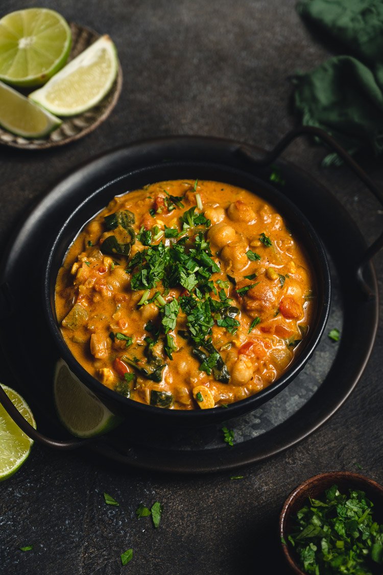 A bowl of chickpea curry garnished with chopped cilantro, surrounded by lime wedges on a dark surface.