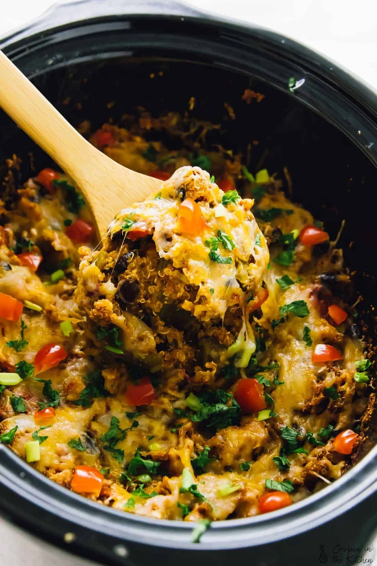 A close-up of a wooden spoon lifting a portion of cooked casserole from a black slow cooker, garnished with chopped green herbs and diced tomatoes.