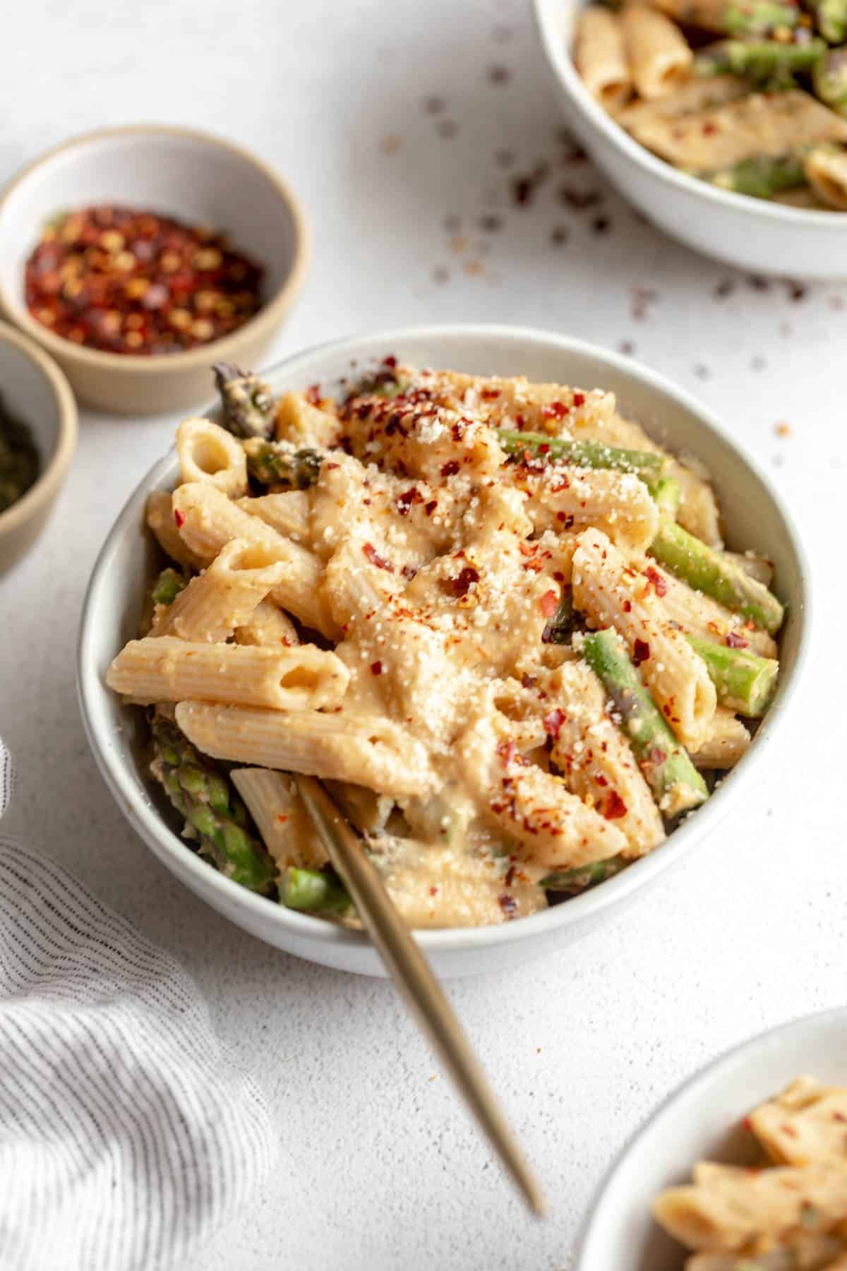 A bowl of creamy pasta with penne, green beans, parmesan cheese, and red pepper flakes, on a white surface with small bowls of chili flakes and herbs.