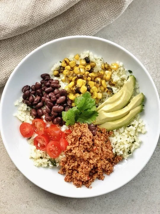 A white bowl with a quinoa base topped with black beans, grilled corn, sliced avocado, cherry tomatoes, meatless crumble and cilantro.