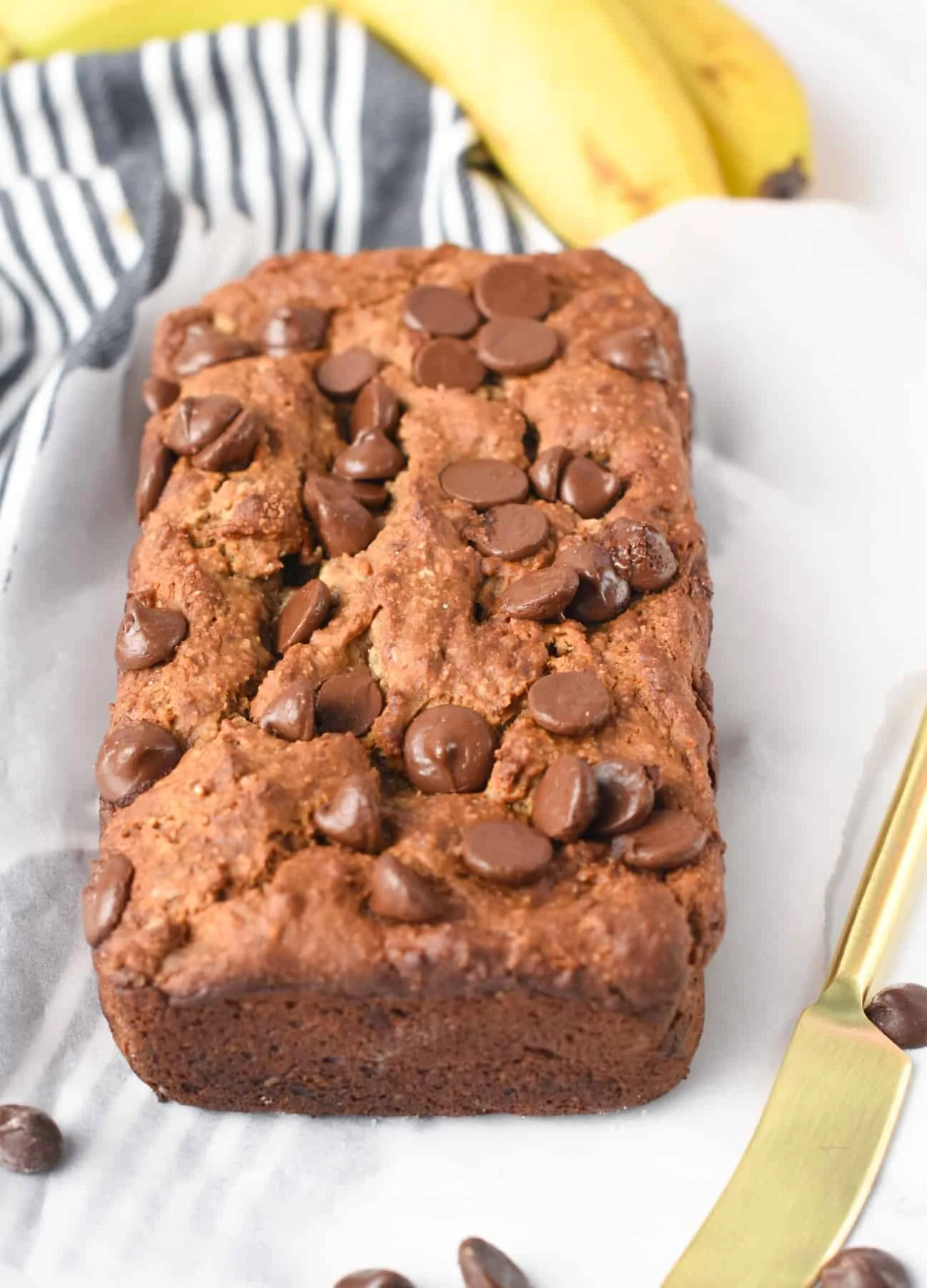 Chocolate chip banana bread loaf on white parchment, with a gold knife and bananas in background.