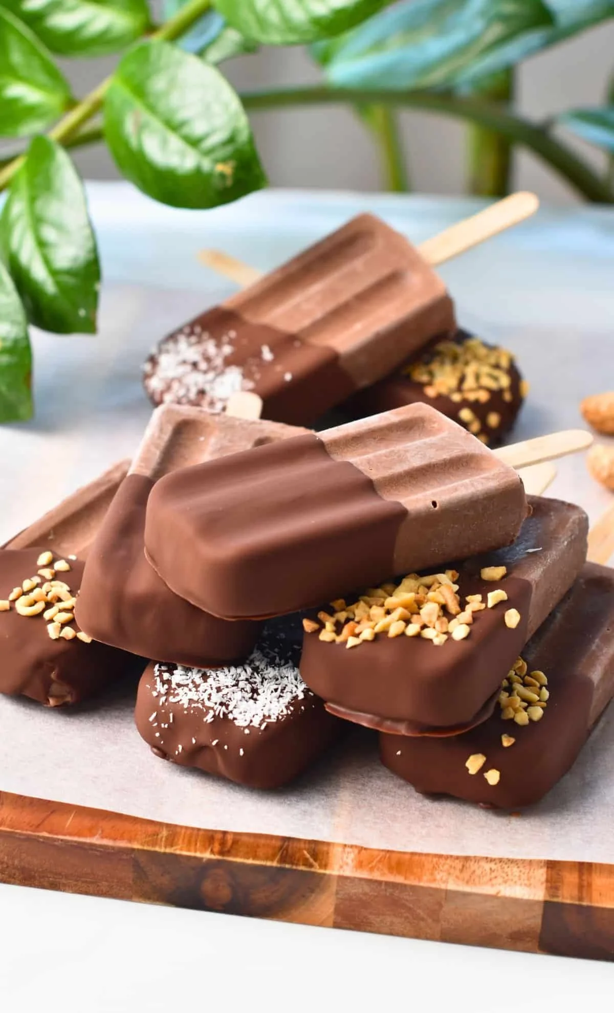 Assorted chocolate popsicles on a wooden tray, garnished with nuts and coconut, with green leaves in the background.