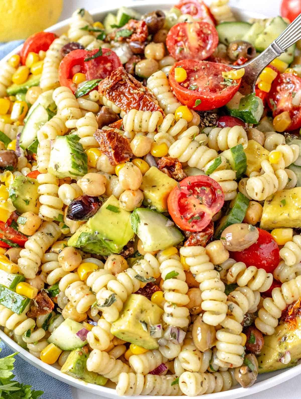 A plate of vegan pasta salad with spiral pasta, cherry tomatoes, cucumber, corn, chickpeas, sun-dried tomatoes, and chopped herbs.