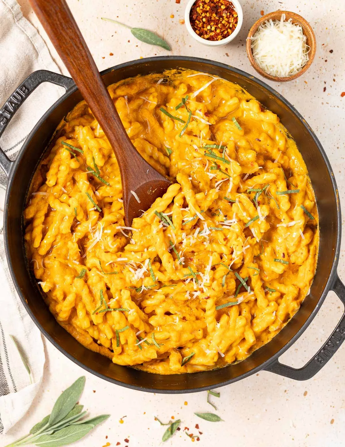 A skillet of cheesy cavatappi pasta garnished with grated cheese and herbs, with small bowls of crushed red pepper and grated cheese nearby, on a speckled countertop.