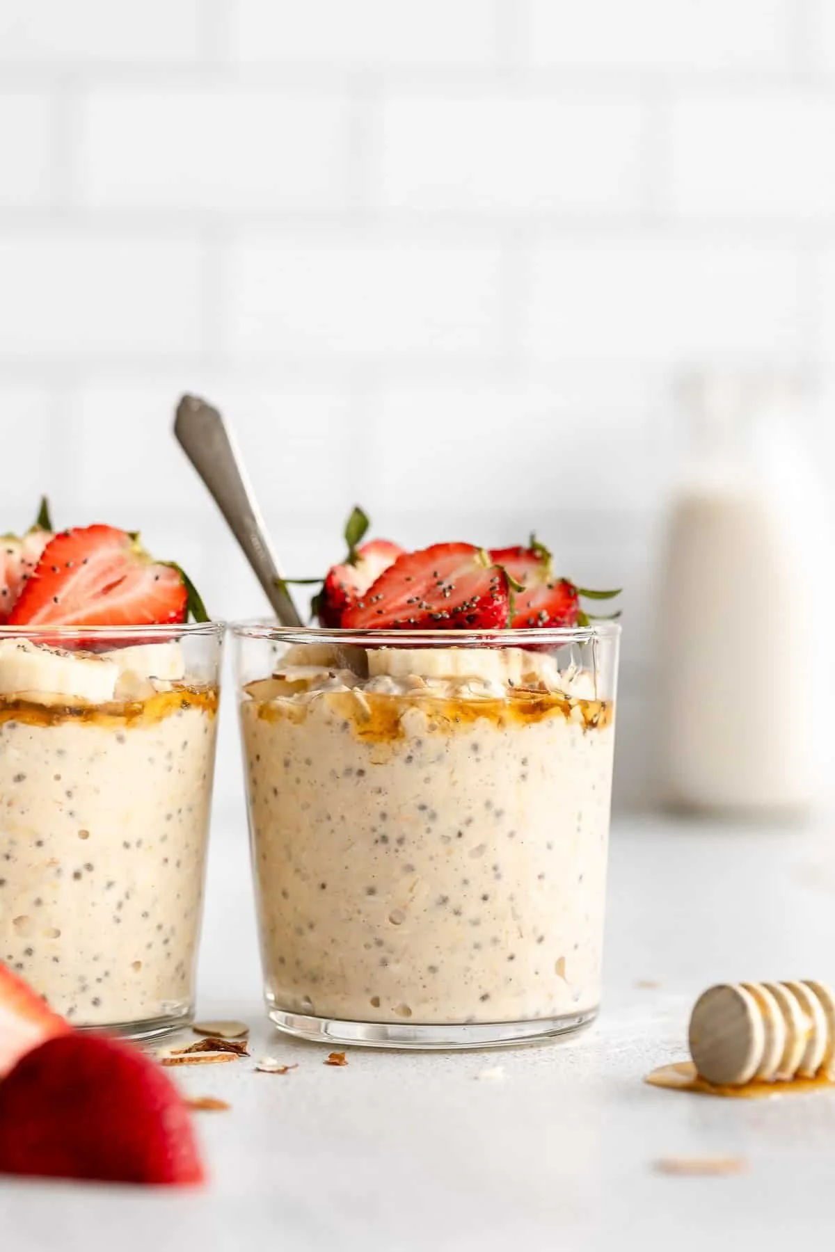 Two glasses of oatmeal pudding topped with strawberries and a drizzle of honey or syrup, with a spoon in one glass and a bottle of milk in the background.
