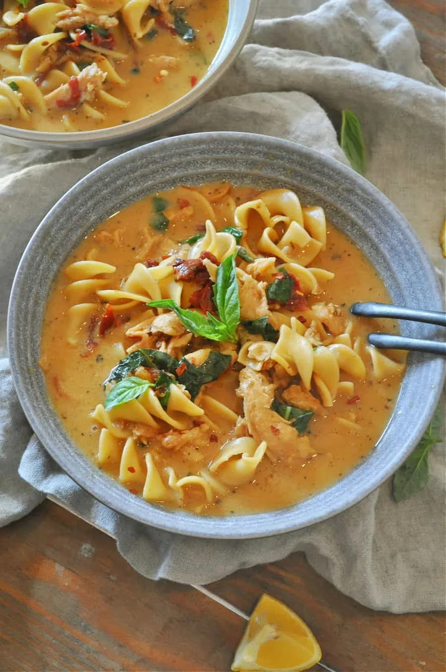 A bowl of vegan noodle soup with pasta, herbs, and vegetables on a linen napkin, with a lemon wedge nearby.