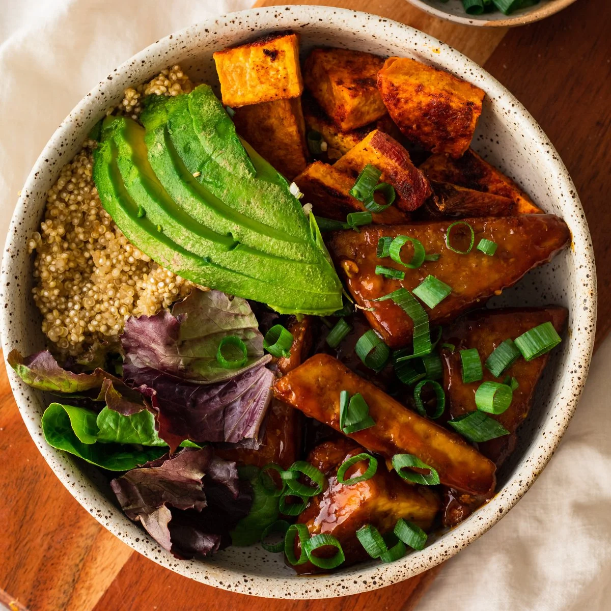 A bowl of cooked quinoa, sliced avocado, roasted sweet potatoes, sautéed chicken in sauce, and mixed greens garnished with chopped green onions.