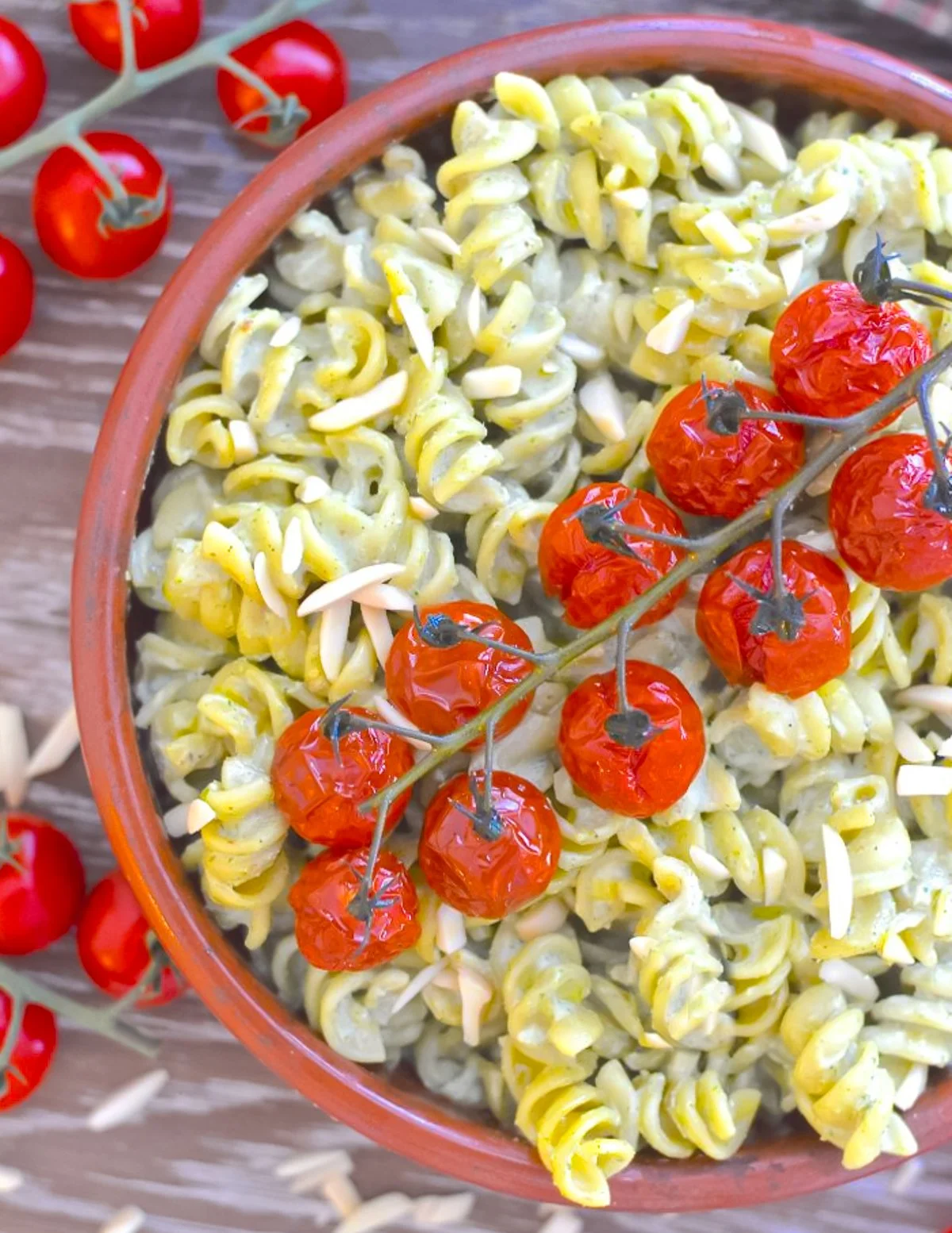 A bowl of pasta salad with fusilli pasta, cherry tomatoes on the vine, and slivered almonds on top, placed on a wooden surface with loose cherry tomatoes nearby.