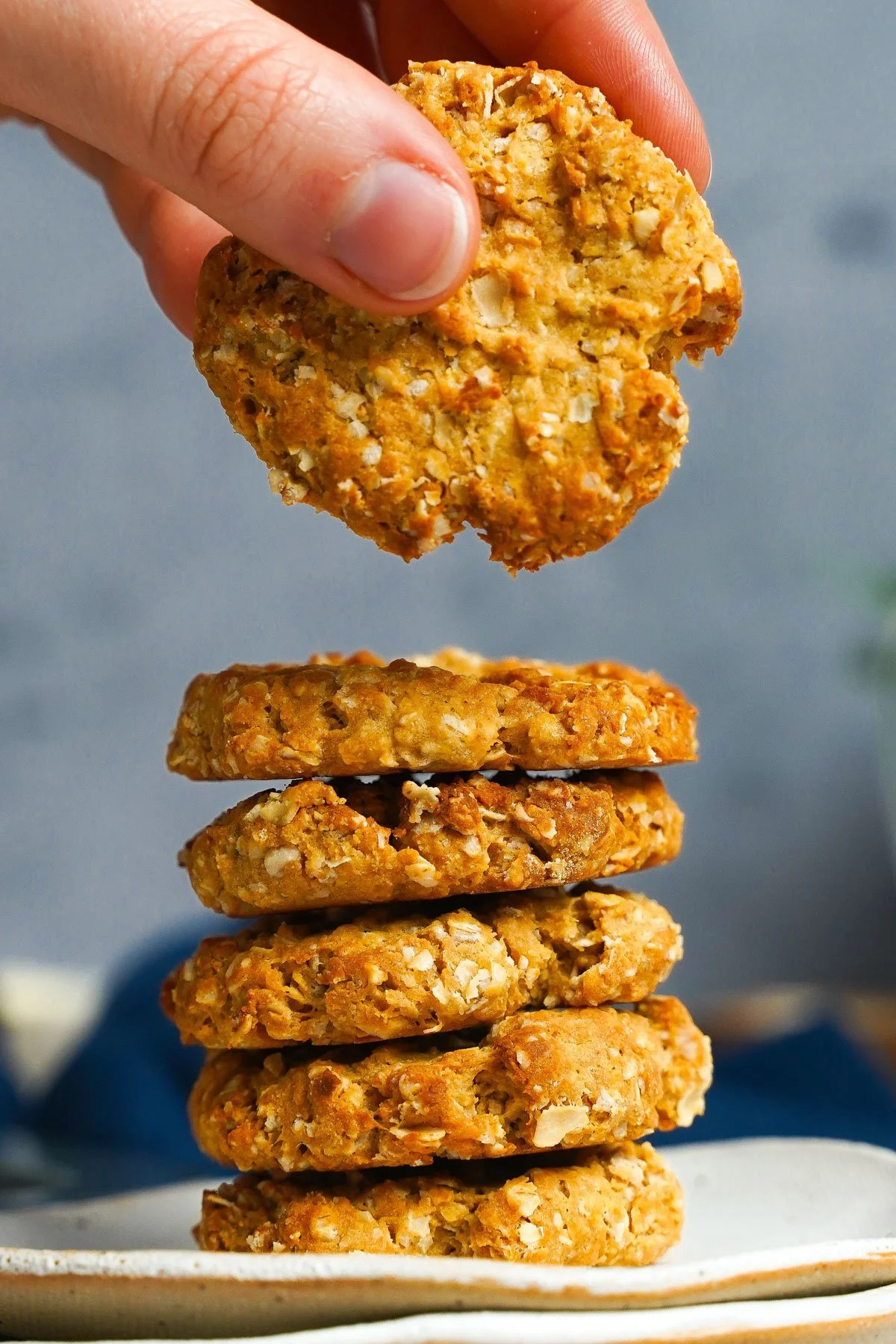 A hand holding one oatmeal cookie above a stack of five similar cookies on a plate.