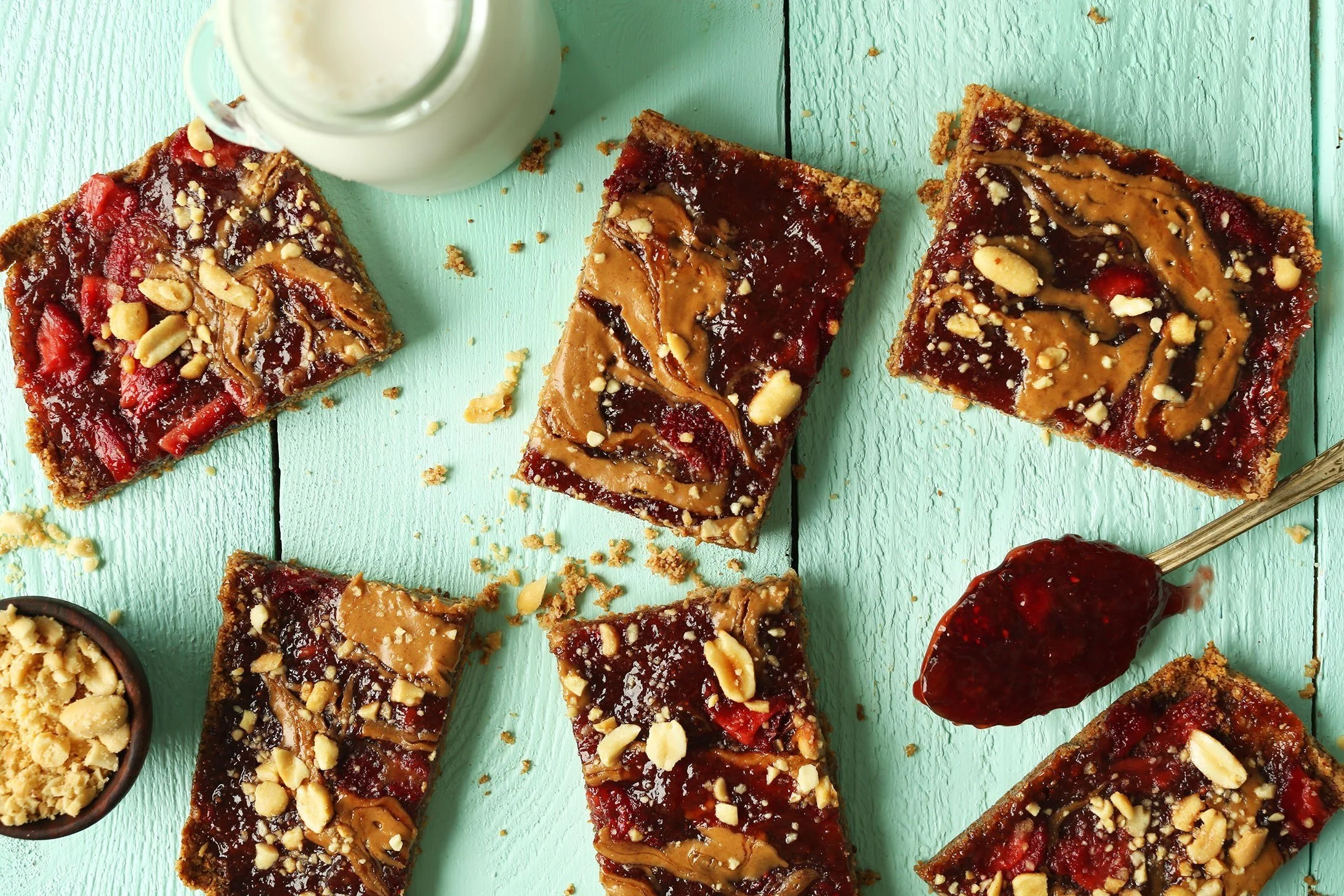 Several slices of kid-friendly vegan peanut butter and jelly dessert bar with chopped nuts on top, arranged on a mint green wooden surface, with a jar of milk and a spoonful of jelly nearby.
