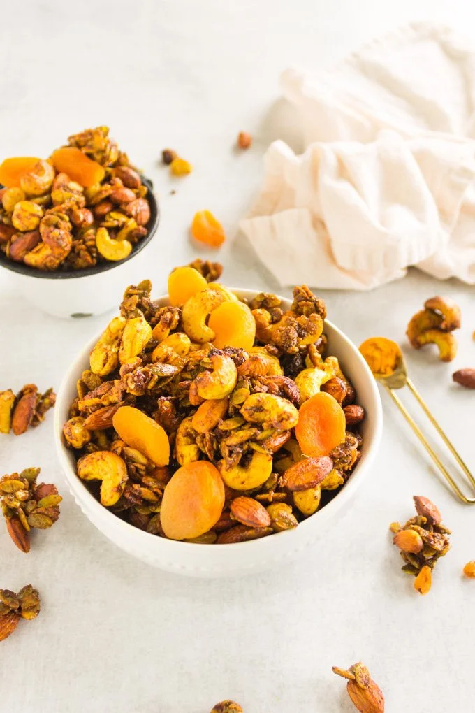 A bowl of homemade nut and dried fruit granola with almonds, cashews, and dried apricots on a white surface, with some scattered granola around and a cloth in the background.