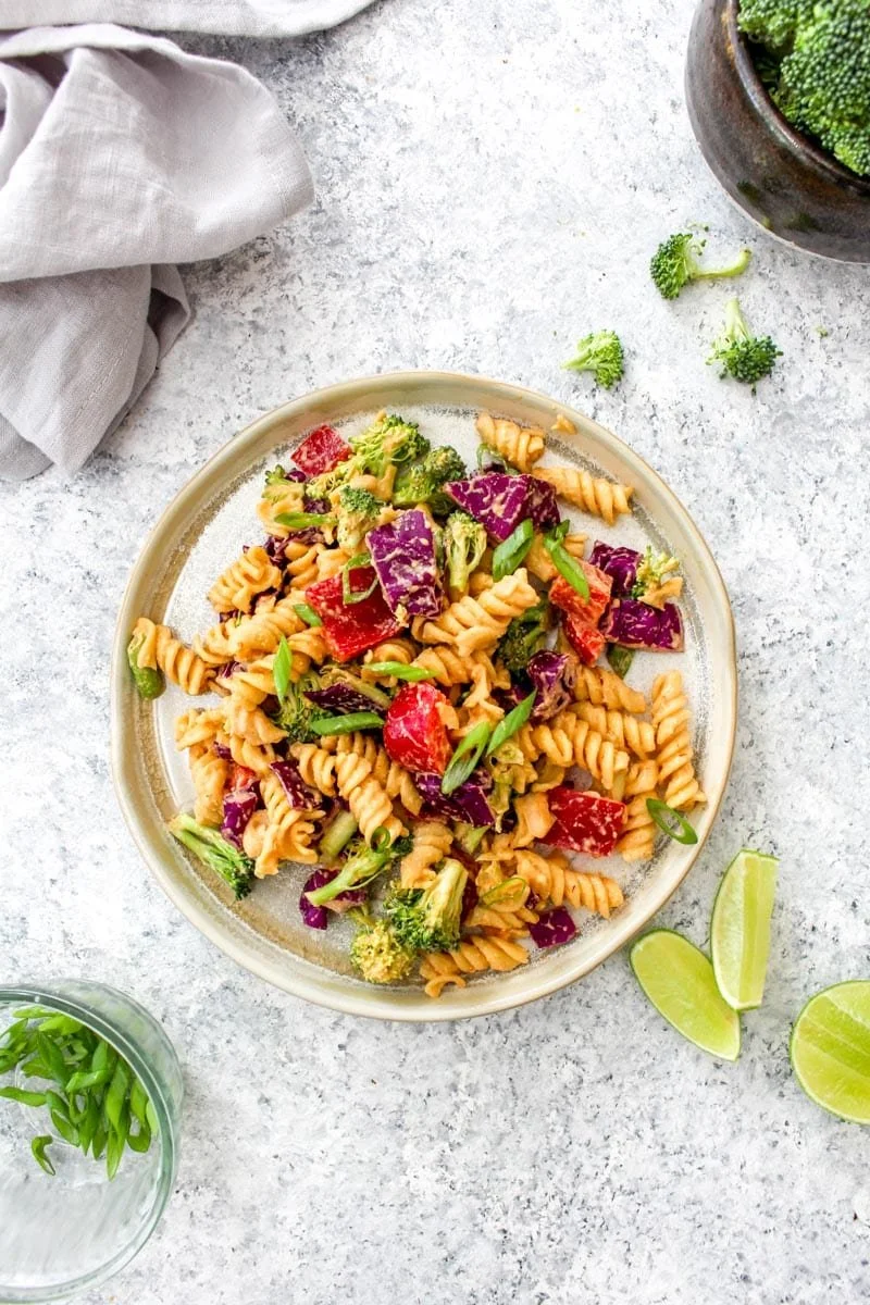 A bowl of spiral pasta salad with broccoli, red bell peppers, purple cabbage, and green onions on a light surface, with lime wedges, a bowl of broccoli, and a glass of water nearby.