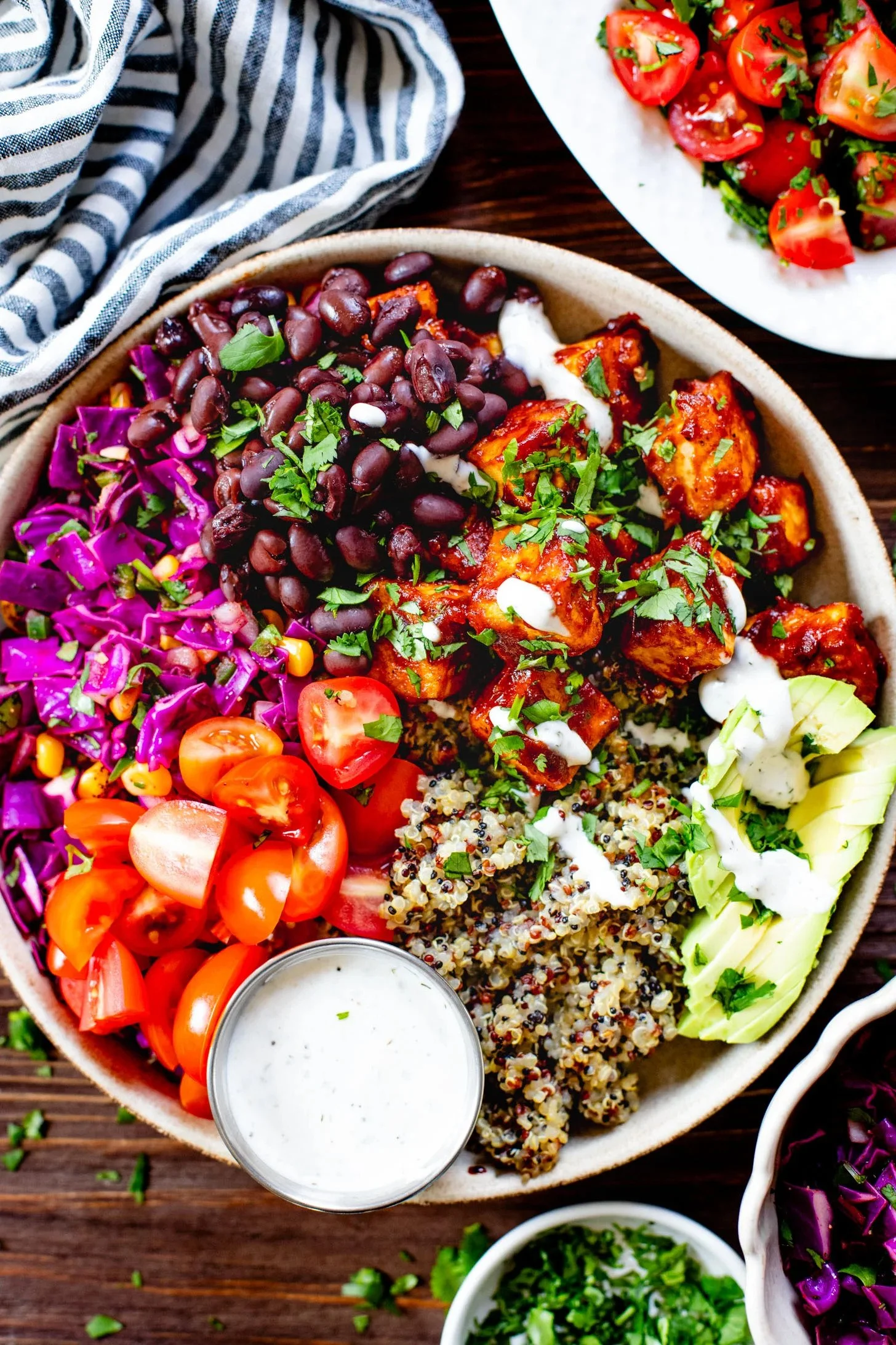 A colorful Mediterranean-style platter with boiled quinoa, cherry tomatoes, sliced avocado, purple cabbage slaw, tofu with sauce, black beans, and creamy dressing, garnished with herbs.