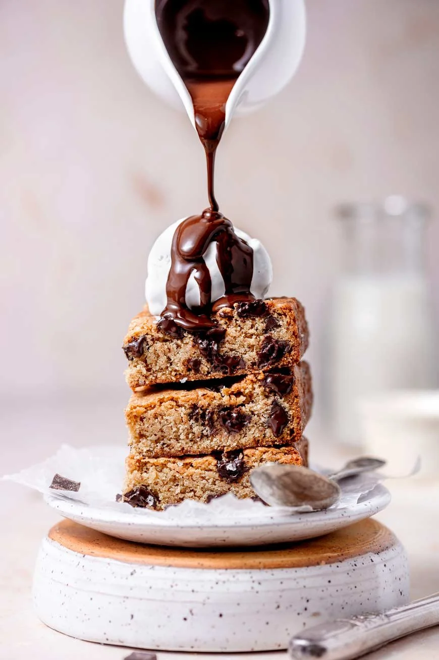 Stack of three chocolate chip cookie bars topped with a scoop of vanilla ice cream, poured with chocolate syrup. A spoon with some chocolate is on the side.