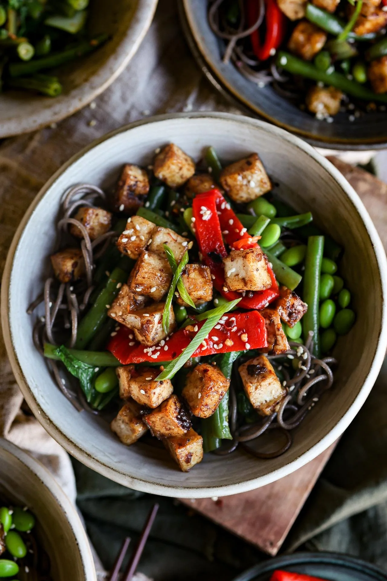 A bowl of Asian-inspired noodles topped with grilled tofu, red bell peppers, green beans, edamame, green onions, sesame seeds, and a drizzle of sauce.
