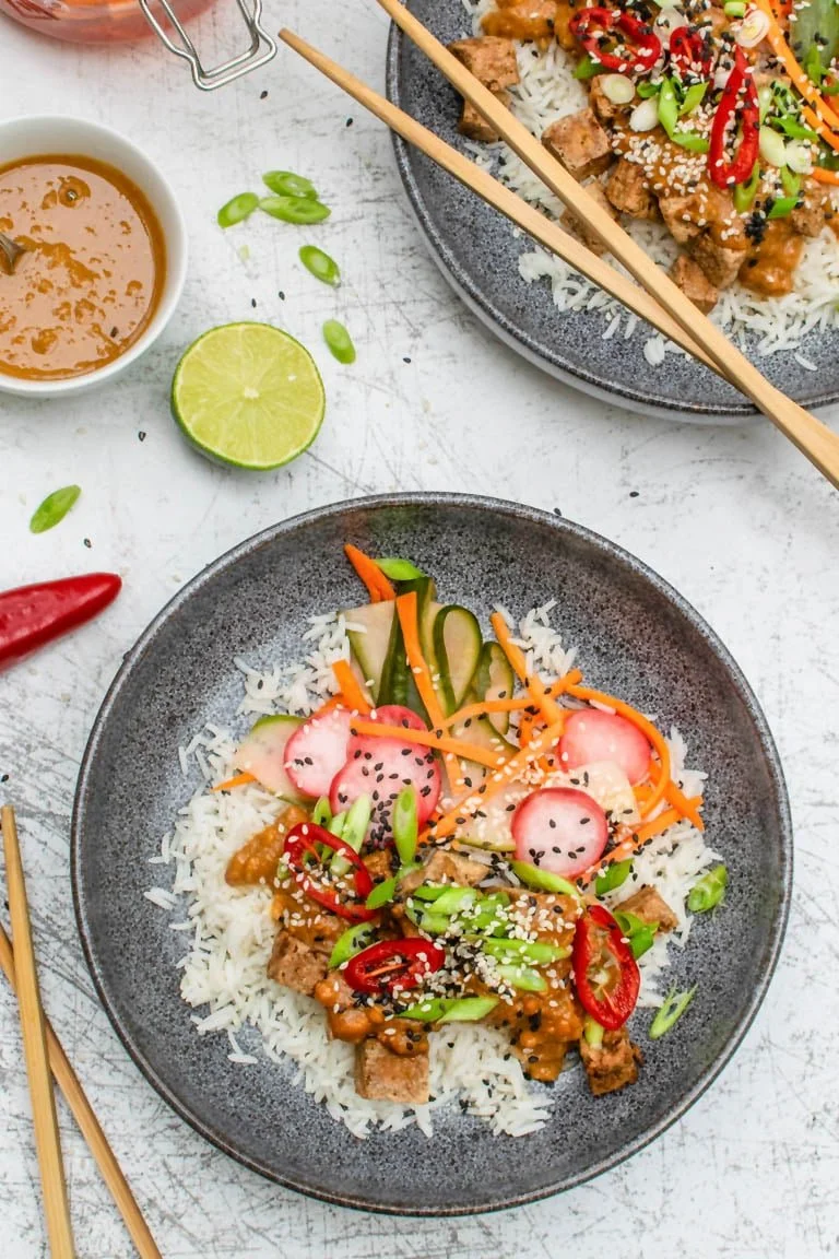 Bowl with rice, vegetables, diced tofu, sliced radishes, sliced red chili peppers, green onions, black sesame seeds, and a sauce, along with chopsticks on a white textured surface, and another similar bowl in the background.
